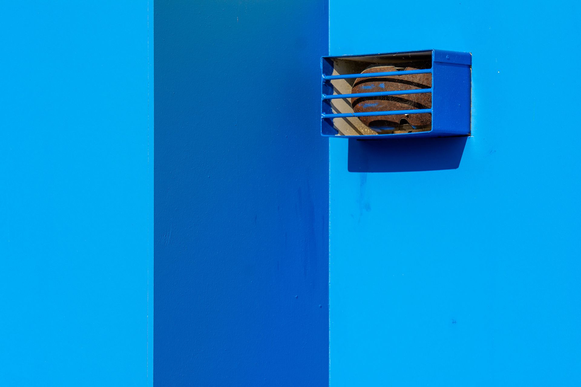 A minimalist shot of a blue wall with rusted vent on sunny day highlighting geometric simplicity.